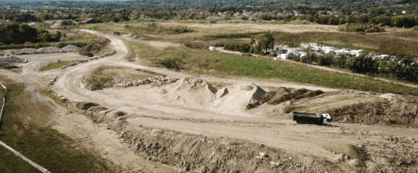 Photo par drone du site actuel côté habitations : pistes, stocks de matériaux et sols nus, avec le quartier voisin en arrière-plan